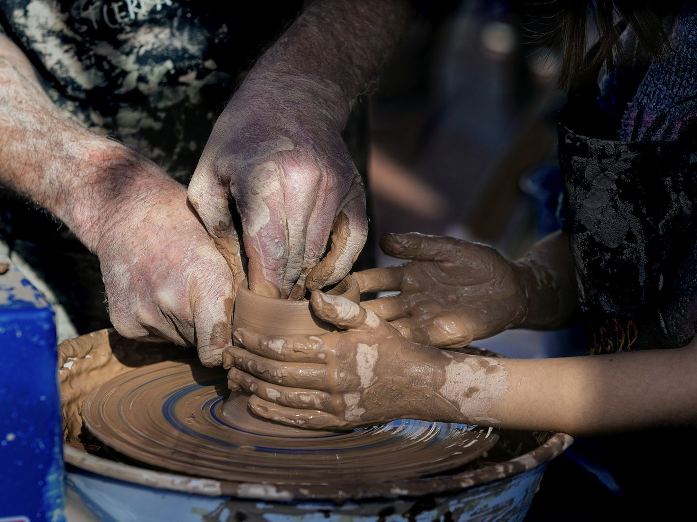 Two pairs of clay-covered hands shaping a pot together on a pottery wheel
