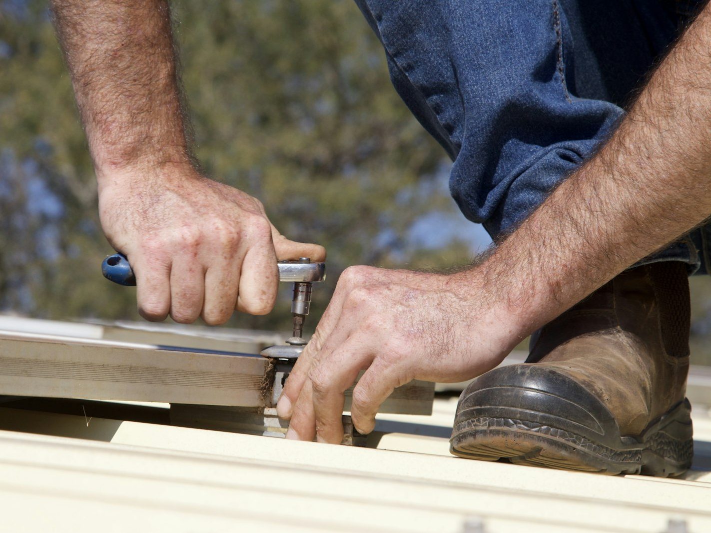 A tradie's hands using a ratchet to fasten corrugated metal roofing, with a work boot alongside